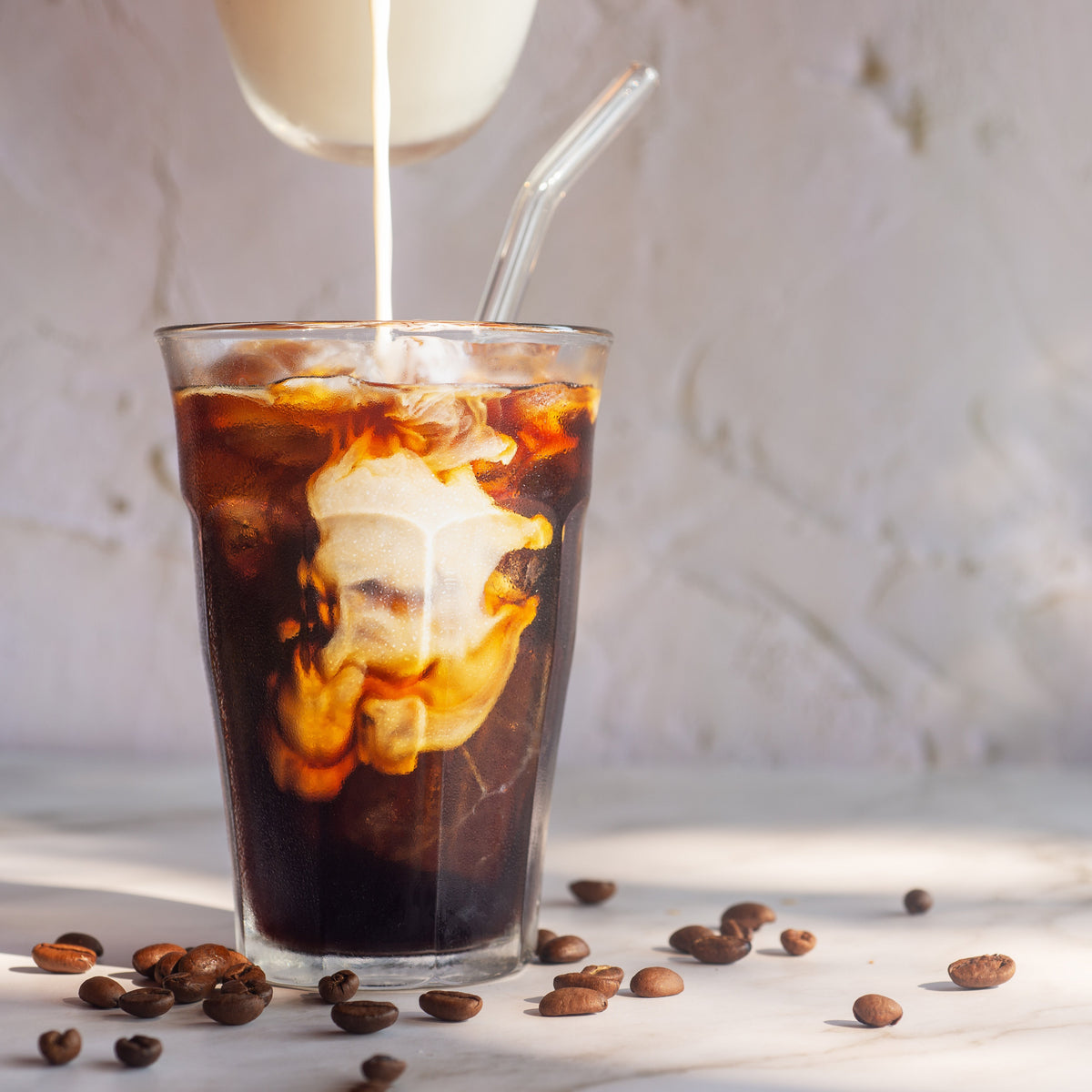 Glass of iced coffee with a straw, being poured into it, on a light surface with scattered coffee beans.
