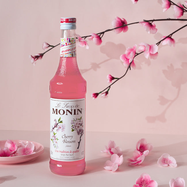 Monin Cherry Blossom syrup bottle with cherry blossom branches and petals on a pink background