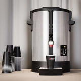 Large coffee urn with digital display and black and white striped cups on a neutral background
