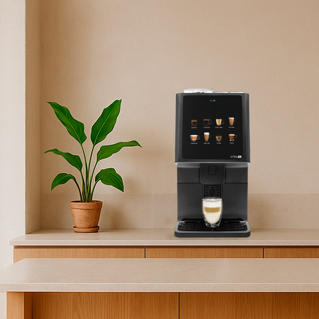A modern black coffee machine sits on a light wood countertop in a warm, minimalist kitchen or cafe setting with wooden stools, a potted plant, and pendant lights.