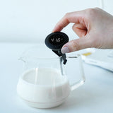 Hand holding a digital thermometer over a glass container of milk with a white background