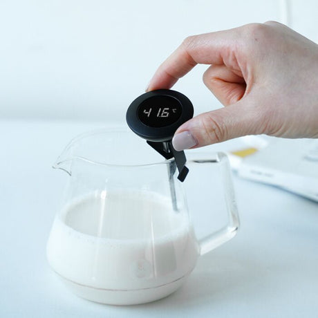 Hand holding a digital thermometer over a glass container of milk with a white background