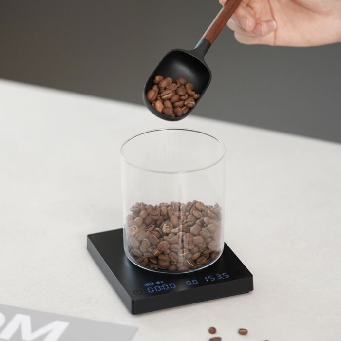 Hand pouring coffee beans into a glass container on a digital scale with a gray background