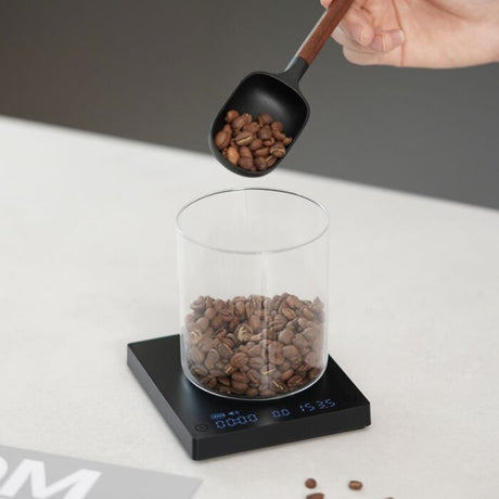 Hand pouring coffee beans into a glass container on a digital scale with a gray background