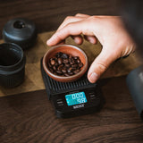 Hand placing a bowl of coffee beans on a digital scale with WACACO branding.