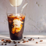 Glass of iced coffee with a straw, being poured into it, on a light surface with scattered coffee beans.
