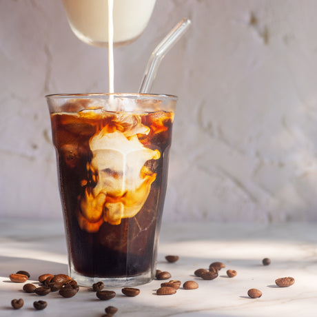 Glass of iced coffee with a straw, being poured into it, on a light surface with scattered coffee beans.