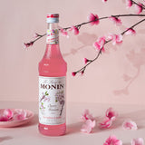 Monin Cherry Blossom syrup bottle with cherry blossom branches and petals on a pink background