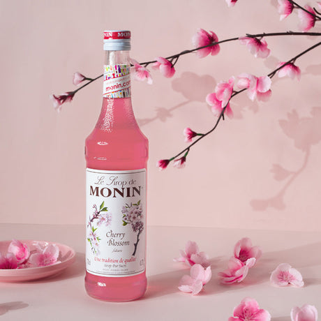Monin Cherry Blossom syrup bottle with cherry blossom branches and petals on a pink background
