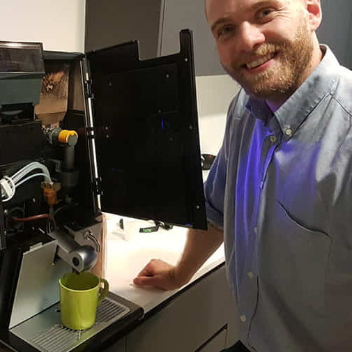 A man in a blue button-down shirt stands next to a compact black countertop water filter machine with its door open, showing the internal components and a green mug placed in the drip tray.